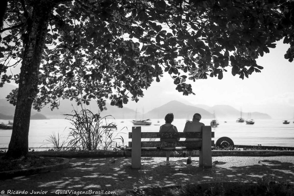Imagem preto e branco de dois idosos sentados em um banco na orla da praia.